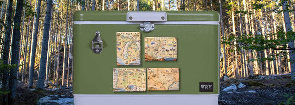 Sitting the woods is a green metal cooler with Michigan, Oregon, Colorado, and Texas magnets on the side. The magnets feature hand-drawn maps by Xplorer Maps.