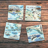 Sitting on a wood table are Xplorer Maps natural stone coasters set of 4. Made from Boccini Marble imported directly from Turkey, showcasing the map of Alaska with intricate details of its vast wilderness, stunning landscapes, and diverse wildlife.