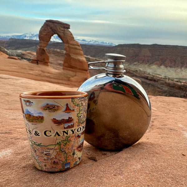 Utah's Arches & Canyonlands National PaArches & Canyonlands National Parks map ceramic shot glass next to a flask. Background: The shot and flask are sitting on a rock shelf overlooking a popular arch rock formation in the National Park.
