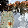 Two people wrapped in a Bryce Canyon fleece blanket next to a snowman. The backdrop is Bryce Canyon National Park red hoodoos..