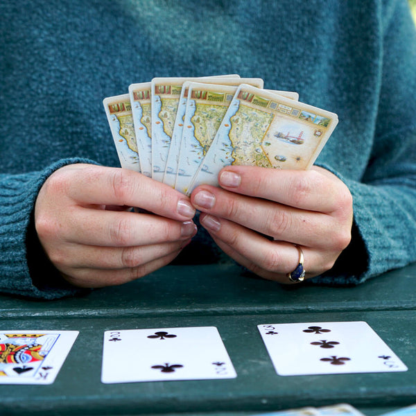 A person playing cards with California State Map on the front. The cards are laying on a green picnic table.