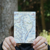 Person holding a Chesapeake Bay wooden postcard in a forest, showcasing hand-drawn map art with regional landmarks and wildlife.