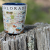 Xplorer Maps shot glass of Colorado sitting on a tree stump. Colorado Map showcases various cities including Denver, Fort Collins, Colorado Springs, Aspen, and Durango. It also features local flora and fauna, such as moose, Rocky Mountain elk, turtles, eagles, and Bighorn Sheep, as well as the indigenous Navajo and Hopi peoples. The map highlights popular activities like hiking, biking, rafting, and skiing.