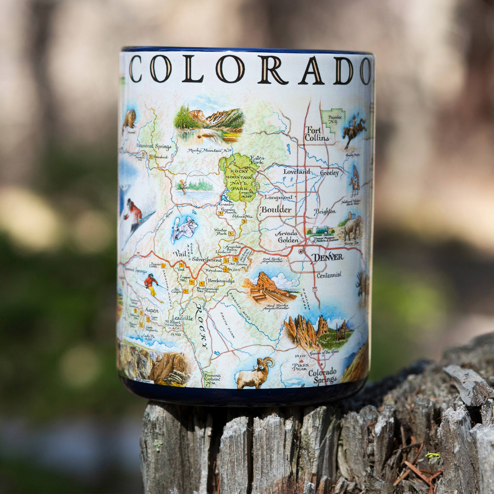 Blue 16 oz Colorado state map coffee mug sitting on a log in the forest. The map showcases various cities including Denver, Fort Collins, Colorado Springs, Aspen, and Durango. It also features local flora and fauna, such as moose, Rocky Mountain elk, turtles, eagles, and Bighorn Sheep, as well as the indigenous Navajo and Hopi peoples. The map highlights popular activities like hiking, biking, rafting, and skiing.