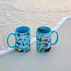Two Blue 16 oz Hawaii Islands Map ceramic mugs on Oahu beach, featuring volcanoes, flowers, ocean, mountains, butterflies, and State Flag