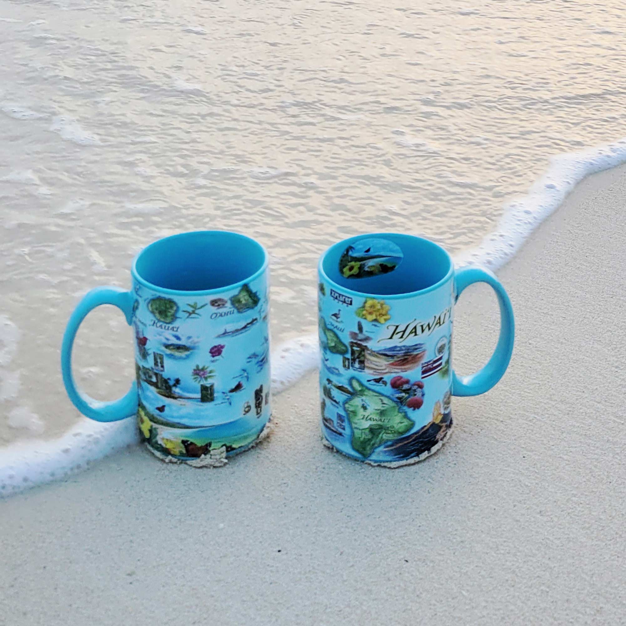 Two Blue 16 oz Hawaii Islands Map ceramic mugs on Oahu beach, featuring volcanoes, flowers, ocean, mountains, butterflies, and State Flag