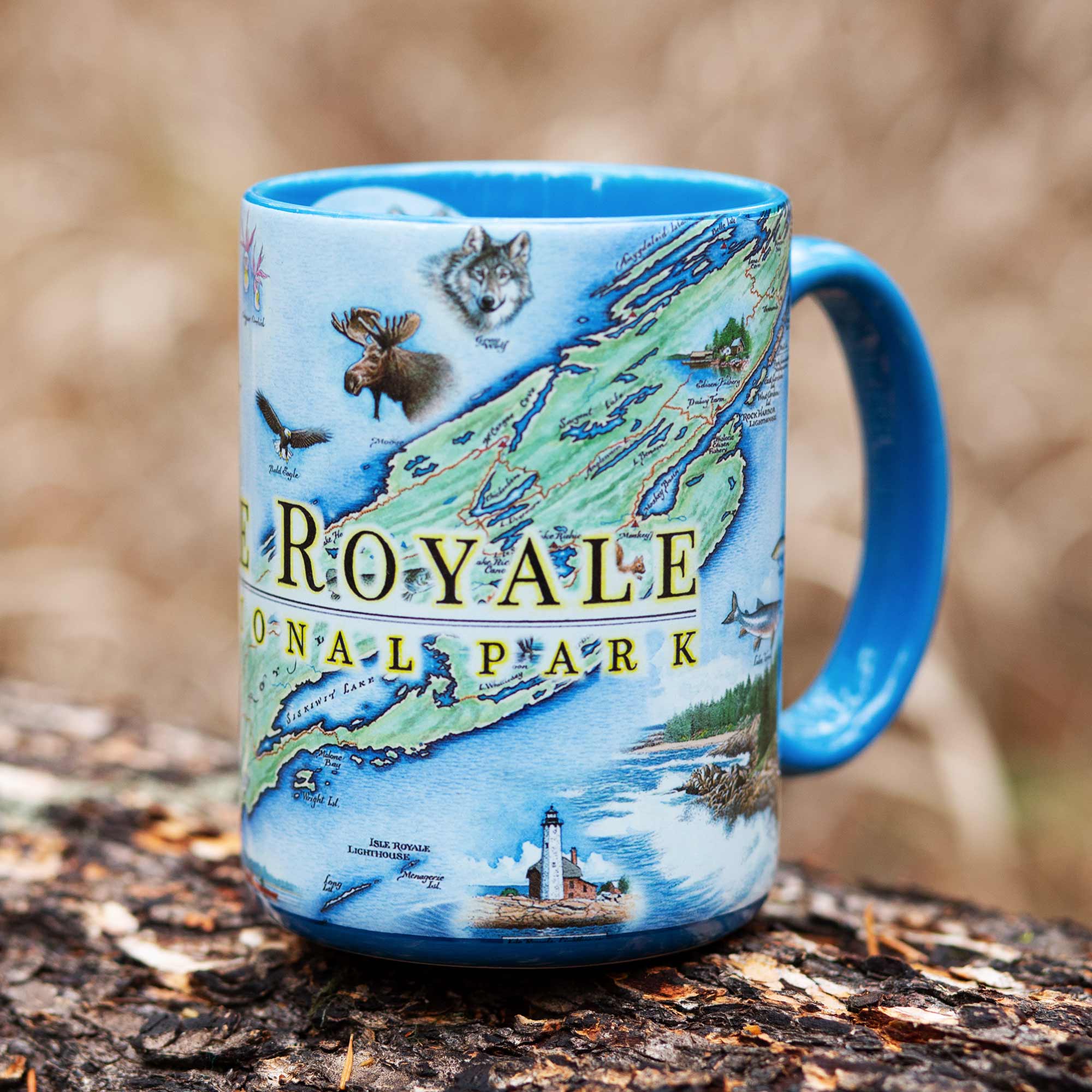 Blue 16 oz Isle Royale National Park Map Ceramic Mug sitting on a log in a forest. The cup features fish, lighthouse, moose, wolf, trees, and lake. 