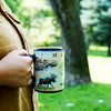 A person holding a Blue, 16oz Maine state map ceramic mug. The background is a tree with green grass. A park like setting.