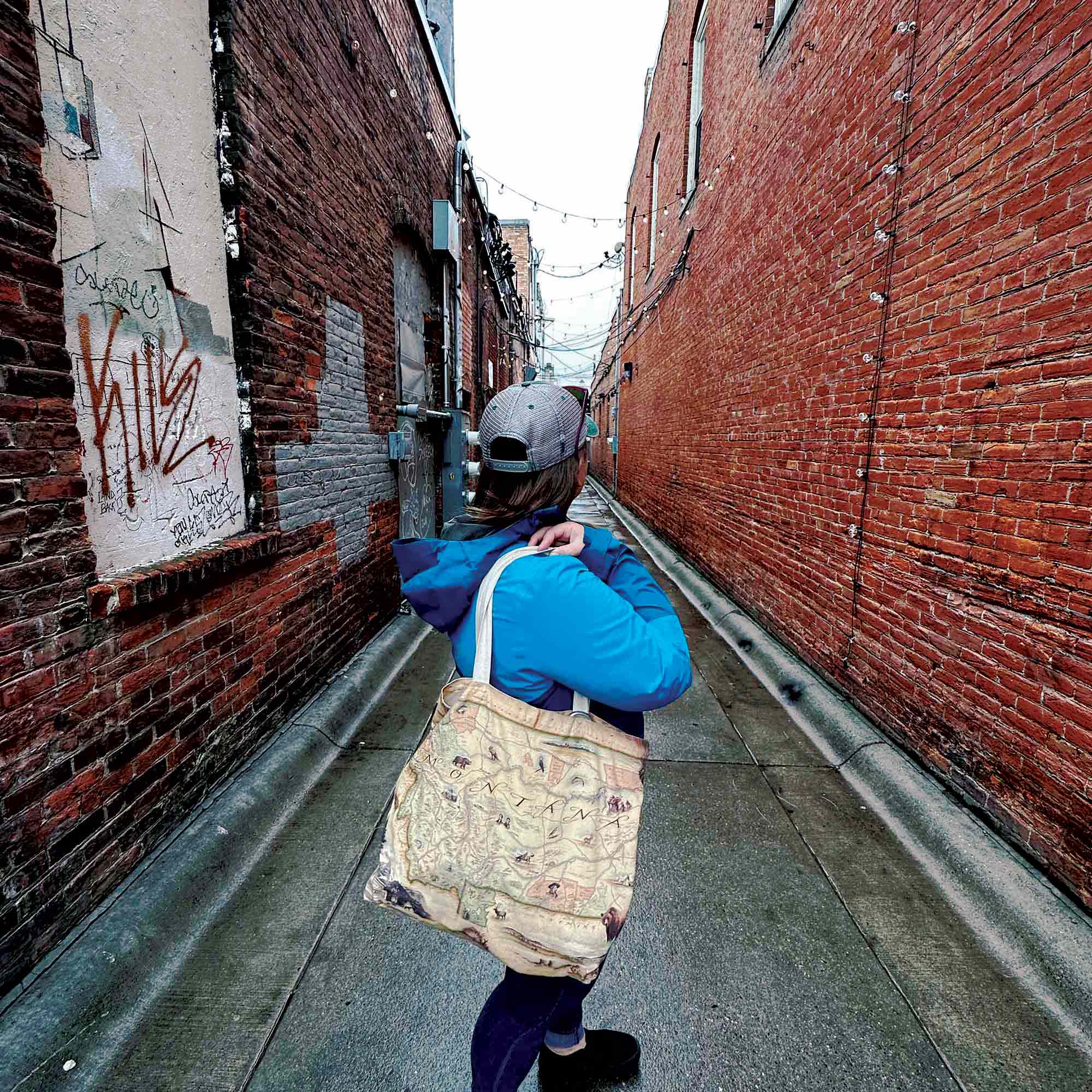 Women in Missoula, Montana alley with Montana Canvas Tote Bag by Xplorer Maps, amidst graffiti walls. Map showcases Sacajawea, Lewis & Clark, Yellowstone, Glacier National Park, Flathead Lake, wildlife like grizzly bear, bald eagle, elk, and cities such as Missoula, Bozeman, Helena, Whitefish.