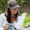 Woman in hat reading 'Fishermans Mercantile Rock Creek, Montana' holds a blue 16 oz ceramic mug of  Rocky Mountain National Park's map. Includes Mount Ganby, Estes Park, Alpine Visitor Center bobcats, snowshoe hares, paintbrushes, and wild roses.