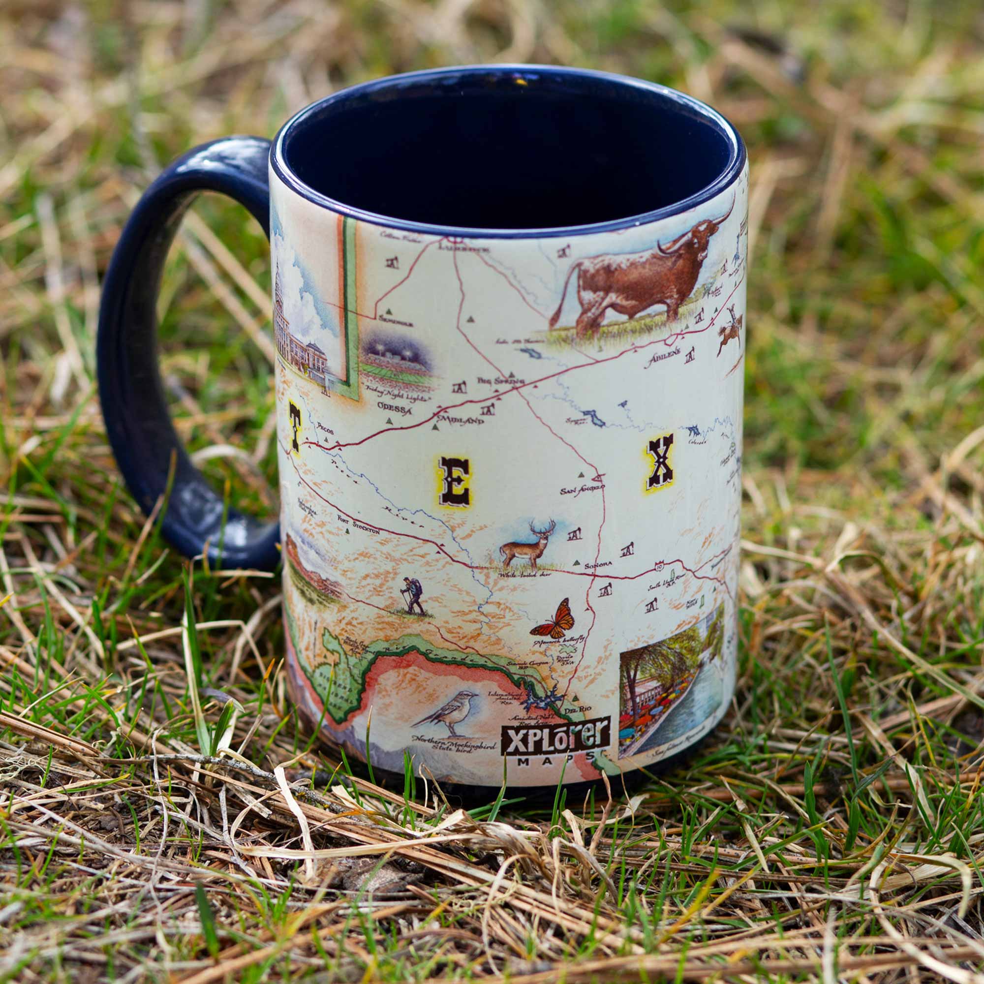 Blue 16 oz Texas State Map Ceramic Mug sitting in grass. The map features illustrations of as the Alamo, San Antonio Riverwalk, and Guadalupe National Park. Flora and fauna include Venus flytraps, longhorns, Monarch butterflies, and armadillos.