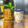 Texas shot glass sitting on a bar next to a tiki cup with pineapple, lime, and mint.