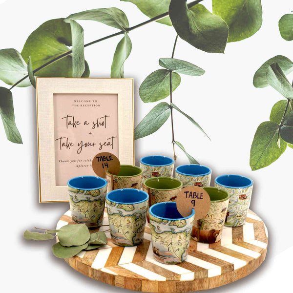 Nine shot glasses sitting on a white and wood  round tray. There is a sign behind it that says, "Take a Shot and Take your seat." The shot glasses have table numbers on a brown tag. 