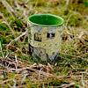 Yosemite National Park Map Ceramic shot glass by Xplorer Maps. The shot is sitting on the grass. Features illustrations of places such as Vernall Falls, El Capitan, and Half Dome. Flora and fauna include mule deer, Indian paintbrush, grey owl, coyote, and a black bear with her cubs. Other illustrations include John Muir, mountain climbing, hiking, and Ansel Adams.
