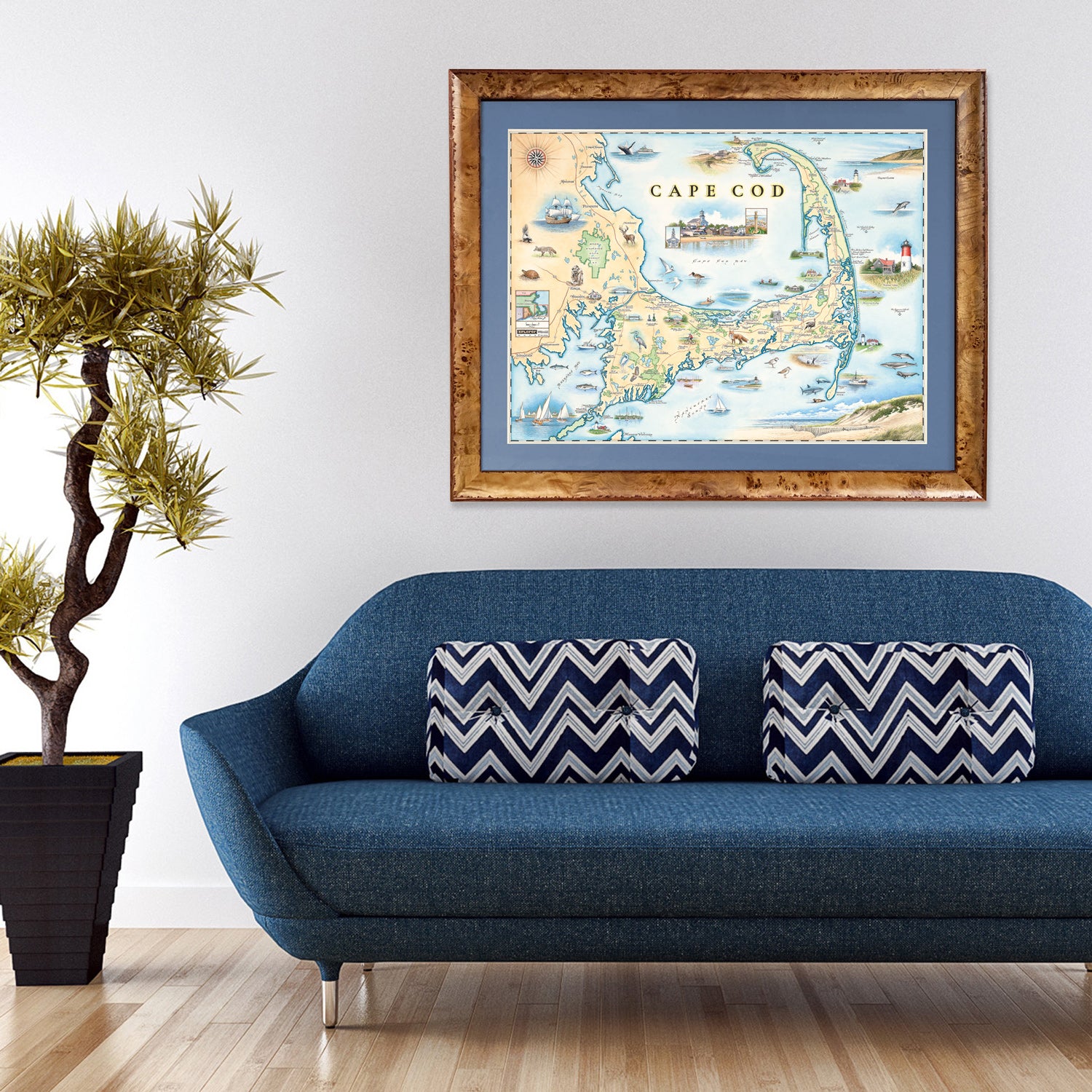 Massachusetts' Cape Cod Hand-Drawn Map hanging over a blue couch.  Featuring Plymouth Rock, fish, crane, fox, Provincetown, canoeing, biking, beach, and ocean.