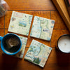 Montana's Flathead Lake Natural Stone Coaster Set of 4 by Xplorer Maps. The coasters are sitting on a tile counter top with a mug full of tea. Features Golfing, Steamboat, Rafting, birds, eagles, Osprey, Bears, forest, water, deer, mountain lion, fish, Cherry Blossoms, and flowers like Glacier Lilies and Lady Slippers. Cities and landmarks are noted such as Woods Bay, Wild Horse Island, Finley Point, and Big Arm.