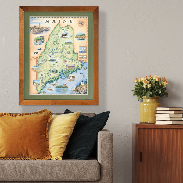 Xplorer Maps state map of Maine hangs in a living room above a gray couch with orange pillows.