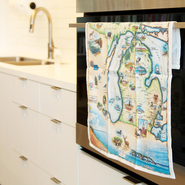 Michigan State Map Kitchen Dishwashing Towel hanging on the kitchen stove handle. The Map features the Great Lakes, Detroit, Ann Arbor, Grand Rapids, and Lansing in earth tones of blue and greens. The tea towel also features Nature, animals, ducks, deer, fish, moose lighthouses, wolverines, and the Mackinac Bridge.