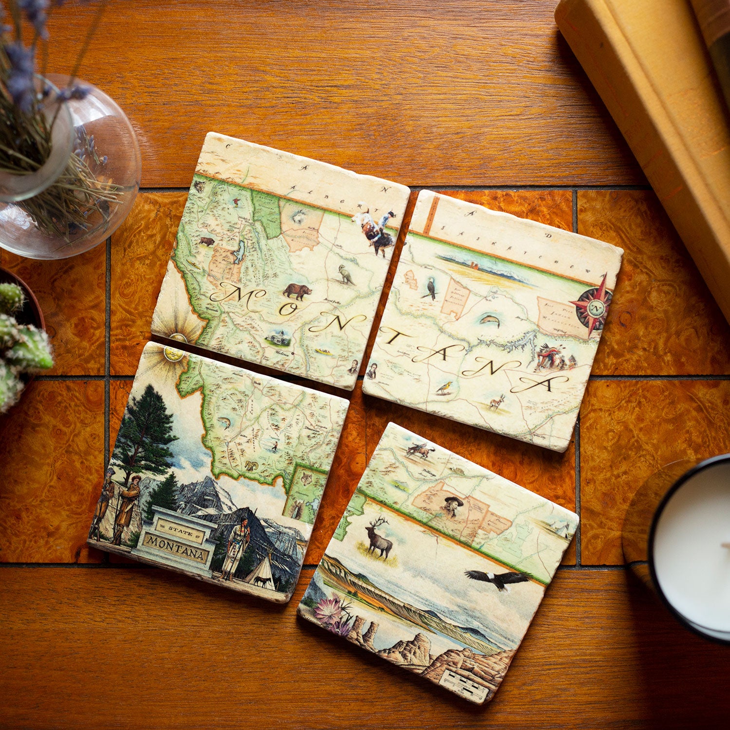 Montana Natural Stone Coaster Set by Xplorer Maps, a set of four coasters forming the map of Montana. Displayed on a tile table with a cactus and candle. The map features iconic landmarks such as Sacajawea, Lewis & Clark, Yellowstone, Glacier National Park, Flathead Lake, and wildlife like a grizzly bear, bald eagle, and elk. Cities including Missoula, Bozeman, Helena, and Whitefish are prominently included in the map