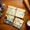 Montana Natural Stone Coaster Set by Xplorer Maps, a set of four coasters forming the map of Montana. Displayed on a tile table with a cactus and candle. The map features iconic landmarks such as Sacajawea, Lewis & Clark, Yellowstone, Glacier National Park, Flathead Lake, and wildlife like a grizzly bear, bald eagle, and elk. Cities including Missoula, Bozeman, Helena, and Whitefish are prominently included in the map