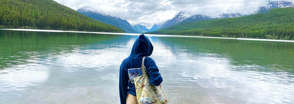 A girl standing at the edge of Glacier Lake in Montana with an Xplorer Maps canvas tote bag. 