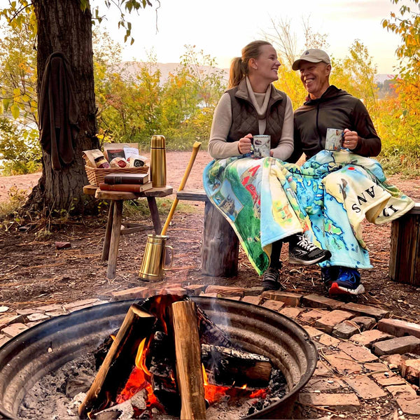 A couple sits by a campfire outdoors, wrapped in a colorful 1000 Islands blanket. The woman and Man holds a mug. Trees, autumn foliage, and a fire pit.