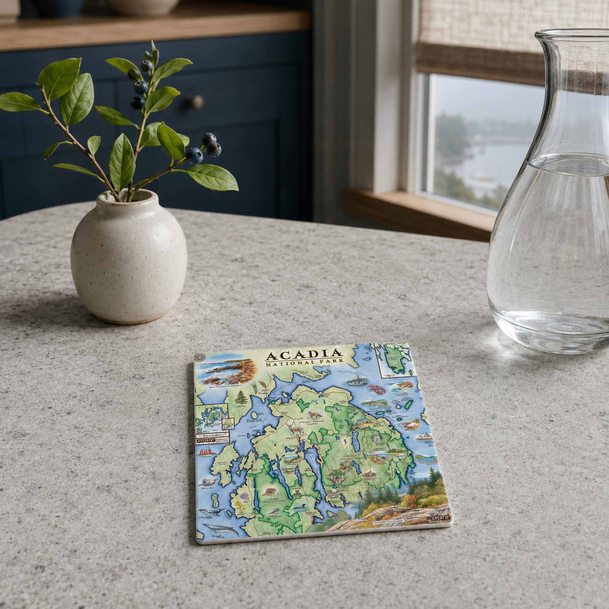 A square ceramic tile coaster featuring a colorful illustrated map of Acadia, styled on a light gray stone countertop next to a small potted plant and a glass carafe.