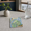A square ceramic tile coaster featuring a colorful illustrated map of Acadia, styled on a light gray stone countertop next to a small potted plant and a glass carafe.