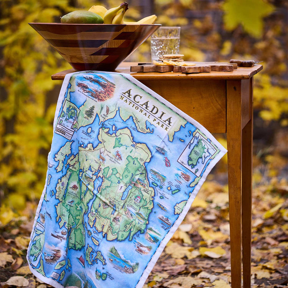 Map of Acadia National Park on a table with a bowl of fruit and glass in an outdoor setting.