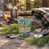 A ceramic mug printed with a colorful map design and the words 'ACADIA NATIONAL PARK' sits on a rustic wooden surface outdoors. Pine branches, stones, a compass, and a plaid blanket are arranged around it, with a blurred campfire and metal pot in the background.