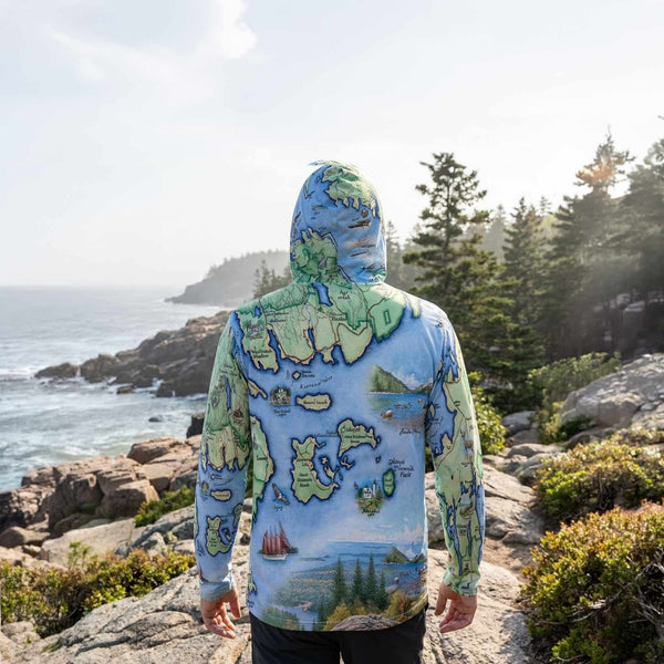 Person wearing a sun shirt hoodie with a map design of Acadia National Park standing on a rocky coastal path.