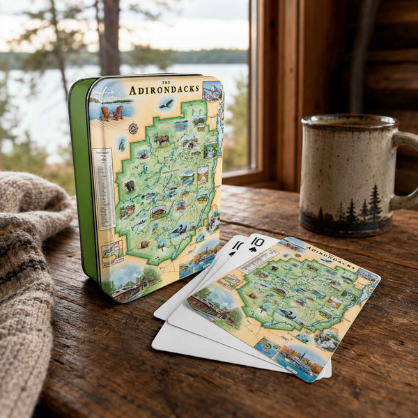A tin and deck of playing cards featuring a vintage-style illustrated map of The Adirondacks, styled on a rustic wooden table by a window with a mug and blanket nearby.