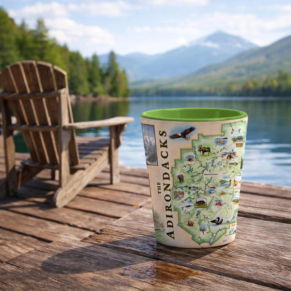 A ceramic travel tumbler featuring a illustrated map of 'The Adirondacks' sits on a wooden dock with a scenic lake and mountains in the background, alongside a wooden Adirondack chair.