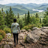 Person standing on a rocky trail wearing Adirondack sun shirt overlooking a scenic landscape with mountains and a lake.