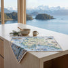 A rectangular printed placemat featuring a vintage-style illustrated map of Alaska, styled on a light stone kitchen island with a ceramic bowl, mug, and evergreen sprig, set against a large window view of snowy mountains and water.