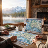 A person and another individual are assembling a large Alaska-themed jigsaw puzzle on a rustic wooden table in a cozy lodge setting. The puzzle box, showing a colorful map of Alaska, sits next to the partially completed puzzle.