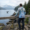 A man stands on a rocky shoreline overlooking a fjord, wearing a light blue hooded long-sleeve shirt with a detailed map and whale print. A humpback whale is visible in the water, with mountains and evergreen forests in the background.