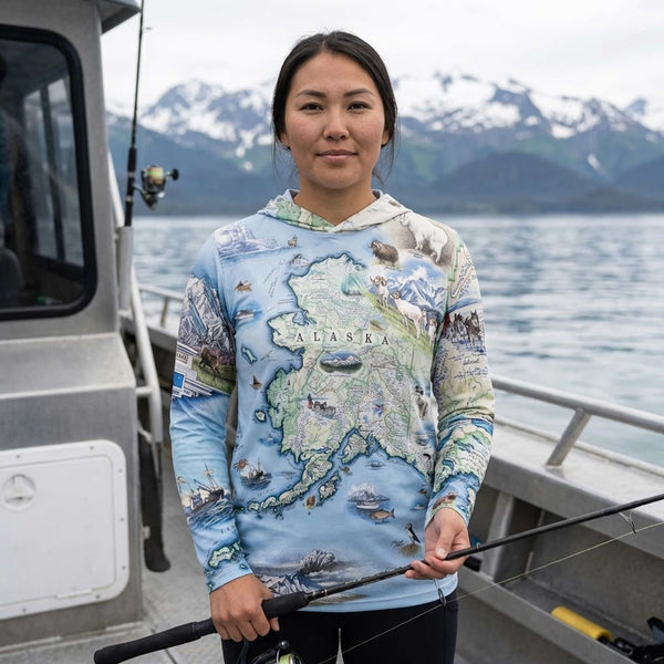 A woman stands on a boat holding a fishing rod, wearing a light blue long-sleeve hoodie with a detailed Alaska map and wildlife print. Snow-capped mountains and water are visible in the background.