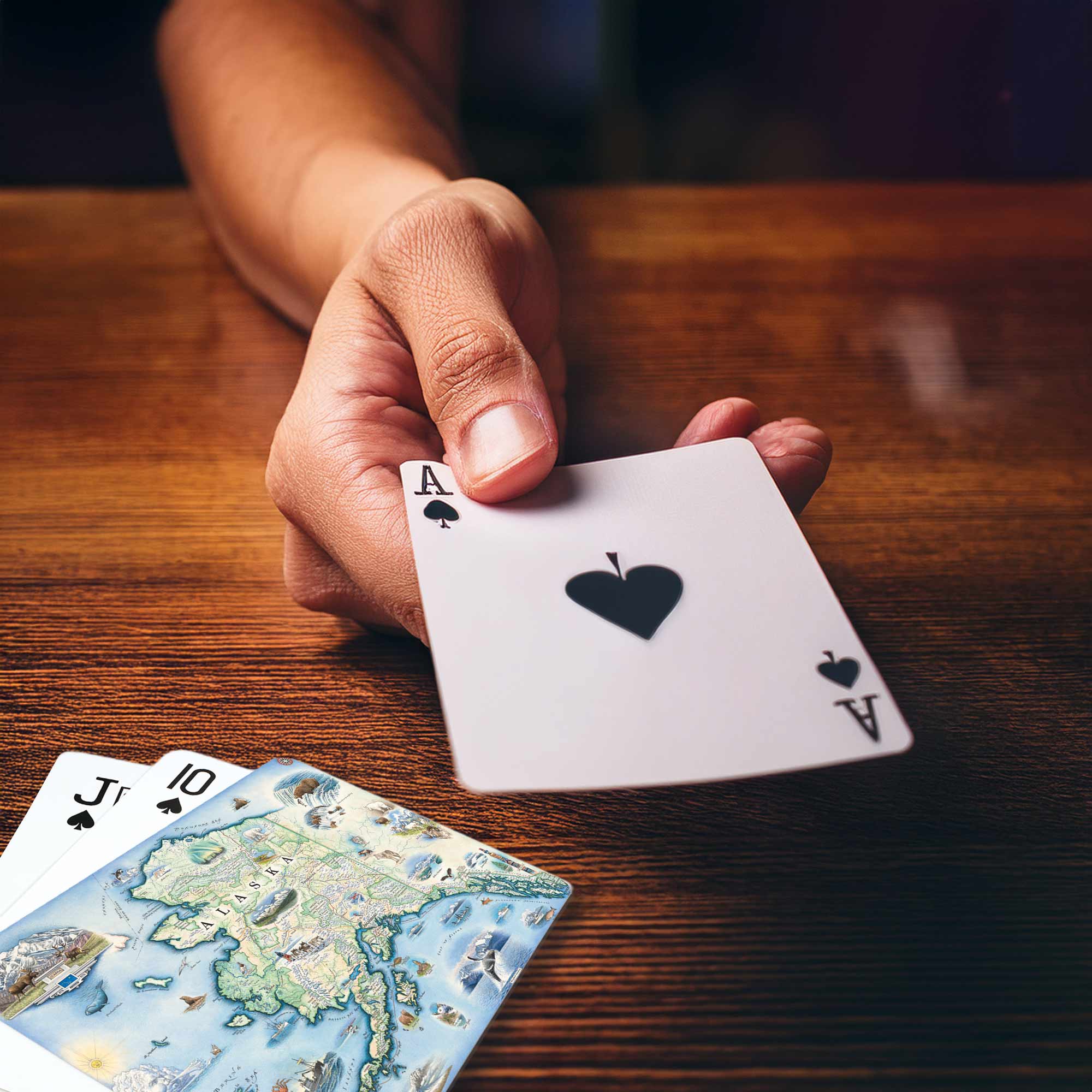 A close-up of a hand holding an Ace of Spades playing card over a wooden table, with two more cards partially visible at the bottom left.
