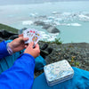 A person outdoors by a glacier or icy landscape holds a fan of playing cards, with a small illustrated metal card tin resting on their blue jacket. The scene includes visible hands, clothing, rocks, and water in the background.