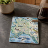 A square ceramic coaster featuring a colorful illustrated map of Alaska with marine and wildlife motifs, displayed on a dark gray stone countertop next to a potted plant and a glass of coffee.