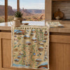 A rectangular woven textile featuring a vintage-style illustrated map labeled 'Arches & Canyonlands' hangs over a wooden kitchen counter. A matching folded textile and a mug sit on the counter, with a real kitchen interior and outdoor canyon view visible in the background.