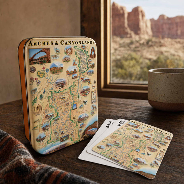 A decorative metal tin and matching playing cards featuring an illustrated map of Arches and Canyonlands, styled on a wooden table near a window with a mug and desert landscape visible outside.