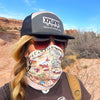 A women in Utah's Arches & Canyonlands National Park stand in front of rock formation with  a gaiter cover her face. 