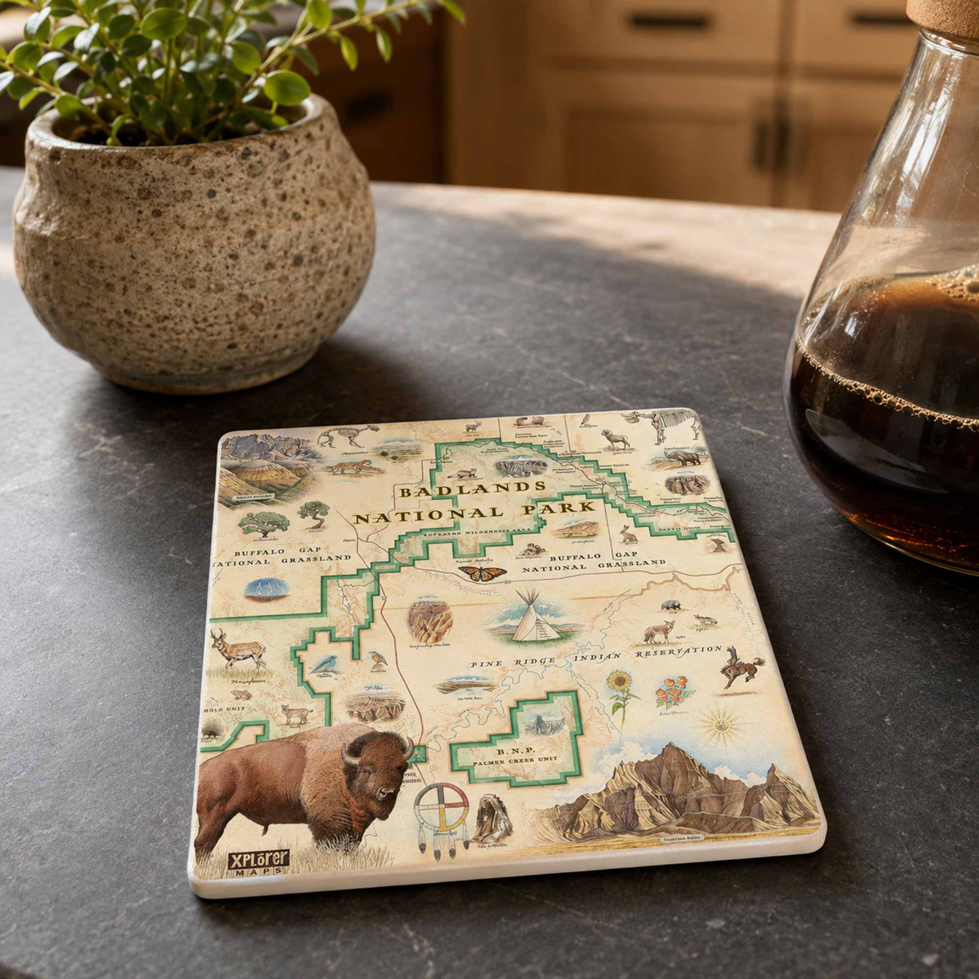 A square ceramic coaster featuring a vintage-style map of Yellowstone National Park with illustrations of a bison, mountains, and green boundary lines, displayed on a dark slate countertop next to a potted plant and a glass bottle of coffee.