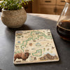 A square ceramic coaster featuring a vintage-style map of Yellowstone National Park with illustrations of a bison, mountains, and green boundary lines, displayed on a dark slate countertop next to a potted plant and a glass bottle of coffee.