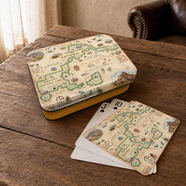A vintage-style illustrated map tin and matching playing cards displayed on a rustic wooden table, with a leather chair and soft indoor light in the background.