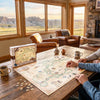 A large illustrated map puzzle and its box are spread on a wooden table in a cozy room with large windows showing a landscape view. Two people are seated at the table, and the scene includes chairs, mugs, and a stone fireplace.