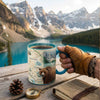 Person holding a mug with a mountain and bear design, set against a scenic lake and mountain backdrop.