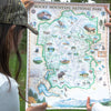 A person holding up a detailed illustrated map of Rocky Mountain National Park outdoors. The map features a green border, topographical line work, and numerous small illustrations of wildlife, lakes, forests, and landmarks.